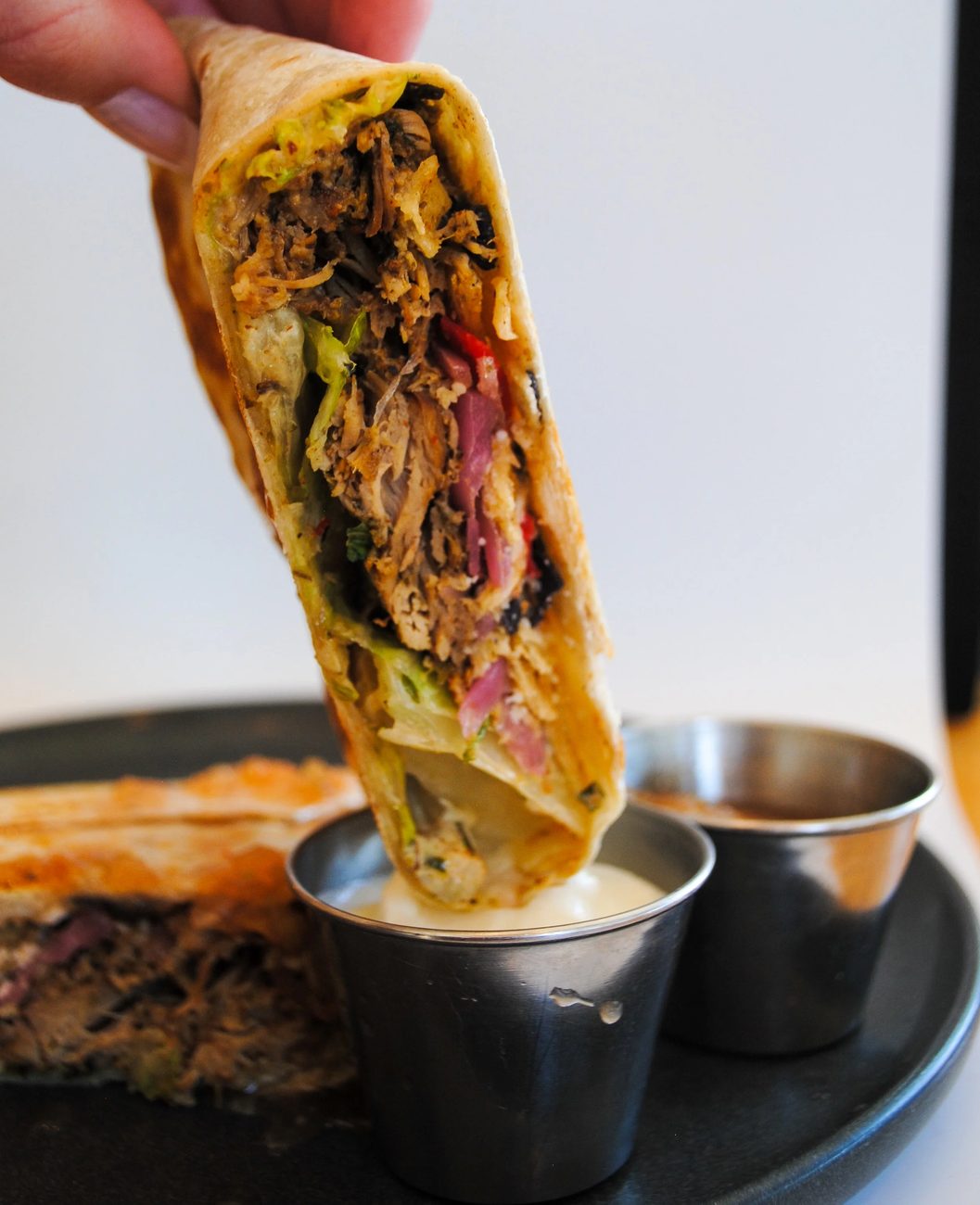 A pulled-pork wrap held over a cup of dipping sauce